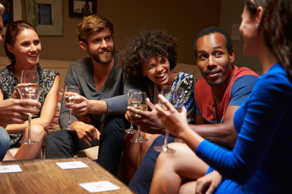 Group of people sitting on a sofa holding glasses of wine; playing cards are on the table in front of them.